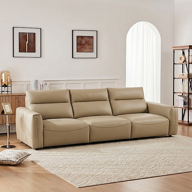 将浅卡其色皮质三人沙发自然融入提供的客厅背景中，移除背景中原有的白色沙发。沙发稳固落地，保持饱满填充的质感，光影与背景匹配：柔和漫射光从右侧拱形窗射入，在沙发底部生成自然的接触阴影，沙发表面反射环境的暖调光线，边缘无抠图白边，与场景融合自然。画面保留背景中原有的装饰画、边柜、书架、地毯等元素，整体为商业家居摄影风格，画面细腻真实。