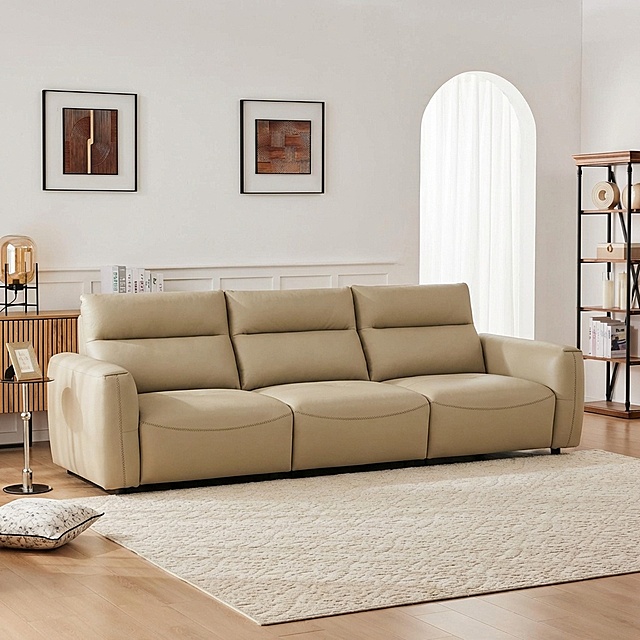 将浅卡其色皮质三人沙发自然融入提供的客厅背景中，移除背景中原有的白色沙发。沙发稳固落地，保持饱满填充的质感，光影与背景匹配：柔和漫射光从右侧拱形窗射入，在沙发底部生成自然的接触阴影，沙发表面反射环境的暖调光线，边缘无抠图白边，与场景融合自然。画面保留背景中原有的装饰画、边柜、书架、地毯等元素，整体为商业家居摄影风格，画面细腻真实。