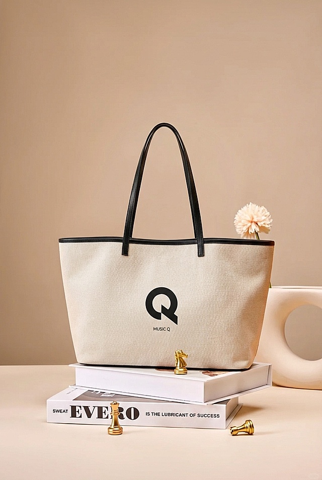 将米白色帆布托特包（带有黑色Q形logo和黑色包边、提手）自然放置在奶咖色桌面场景的堆叠书籍上，完全移除原有棕色包包，完整保留书籍、金色象棋棋子、白色异形花瓶和浅粉花朵。光影匹配场景暖调柔和漫射光，包身有自然明暗层次，底部生成真实柔和的接触阴影，包身边缘融合环境暖棕漫反射，无抠图白边，包的大小比例适配场景，摆放角度自然，整体为高级法式奶油风商业摄影，画面细腻真实，融合自然。