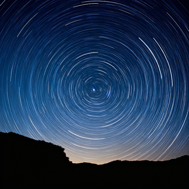 星空轨迹环绕天极，剪影山峦静默守望，粒子光轨交织梦幻夜空，静谧神秘中透出宇宙的深邃律动。