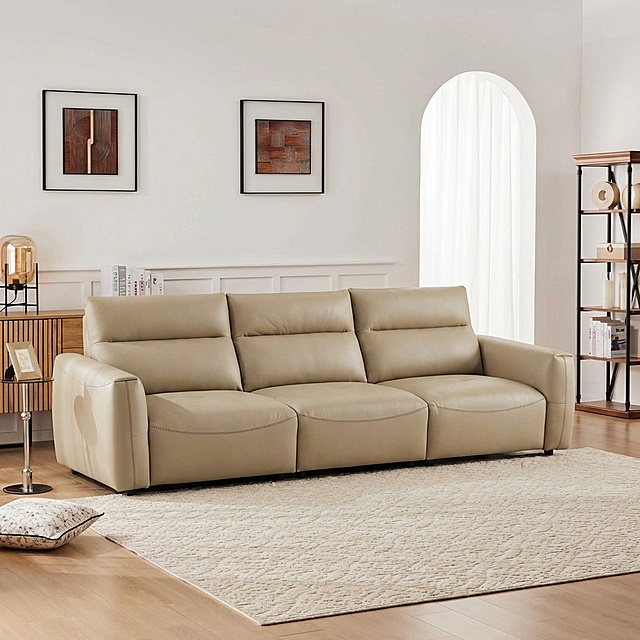 将浅卡其色皮质三人沙发自然融入提供的客厅背景中，移除背景中原有的白色L型沙发。沙发稳固落地，保持饱满填充的皮质质感，光影与背景完全匹配：柔和漫射光从右侧拱形窗射入，在沙发底部生成自然的接触阴影，沙发表面反射环境的暖调光线，边缘无抠图白边，与场景融合自然。画面保留背景中原有的装饰画、边柜、书架、地毯、小边桌、抱枕等所有元素，整体为真实商业家居摄影风格，画面细腻有质感。